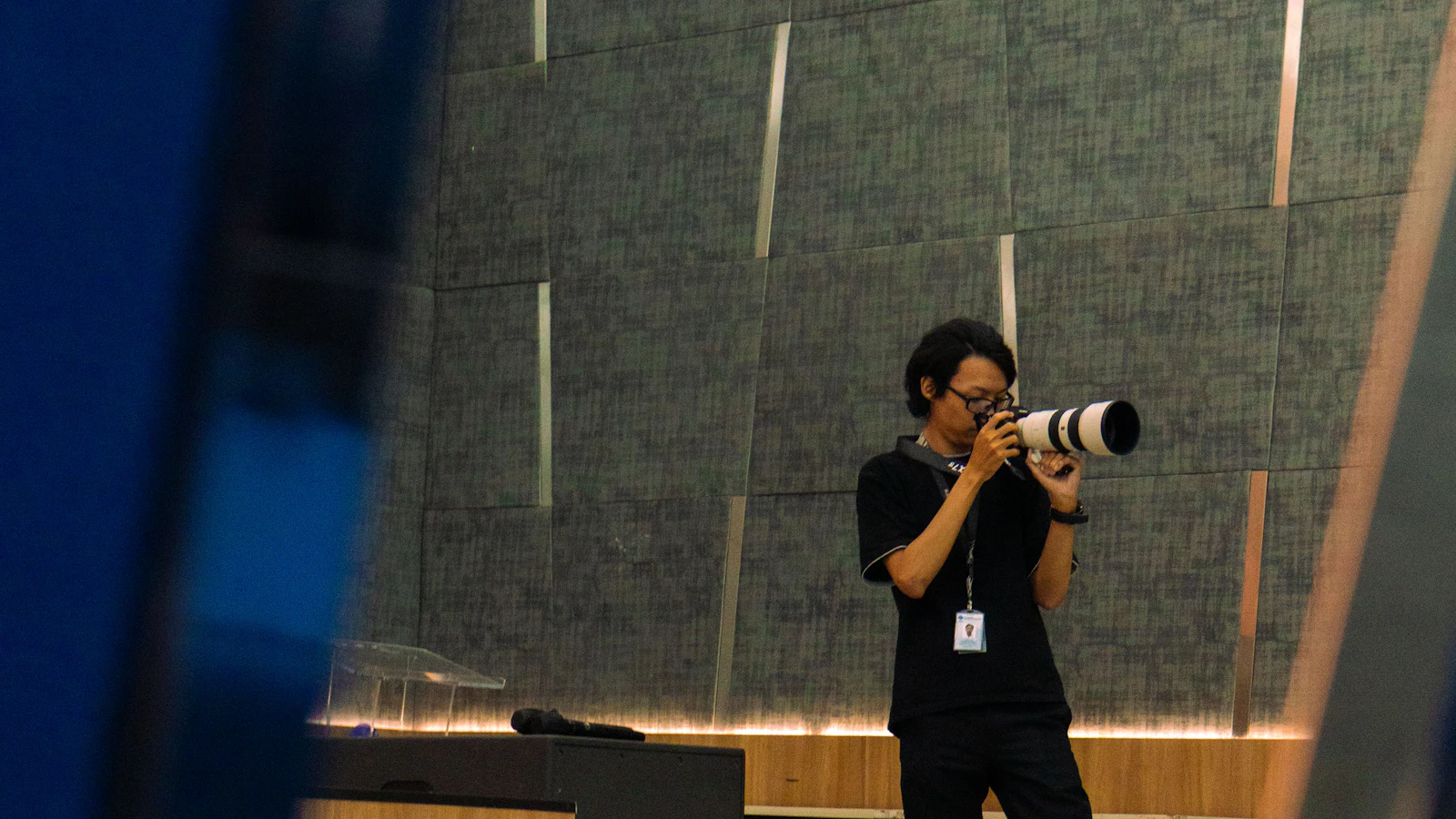 Un photographe au téléobjectif, badge autour du cou, dans une salle de conférence